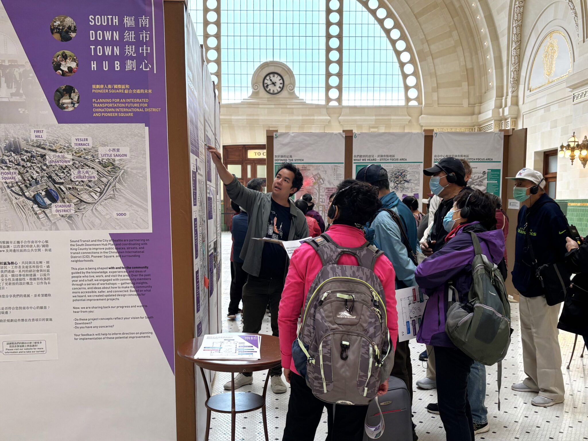 A person points at a display board as a group of elders listen. Some of the elders are wearing backpacks, hats, and face coverings. All of the elders are wearing headsets to listen to language interpretation. On the upper left the display says "South Downtown Hub".