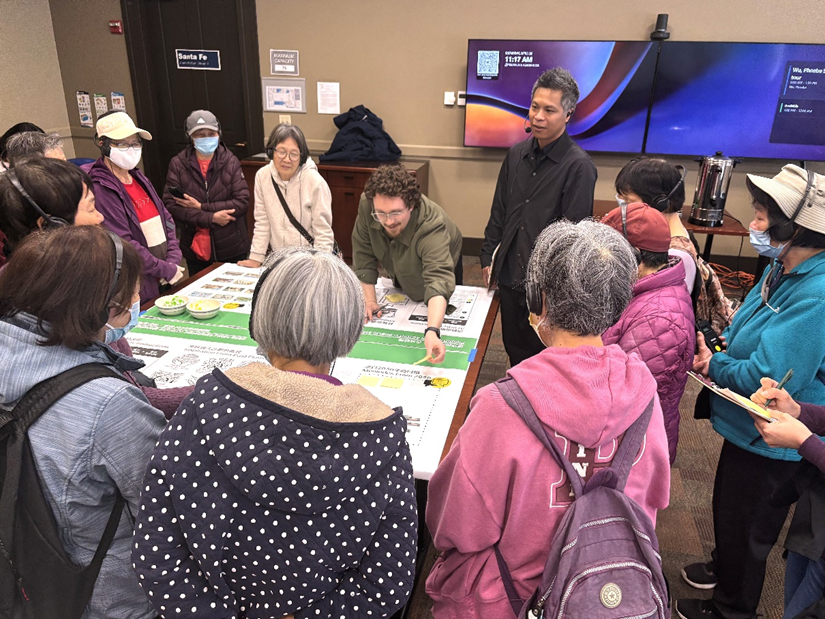 A group of people surround a table that is covered with a large informational poster. Most of the people are elders who are wearing headsets for language interpretation.