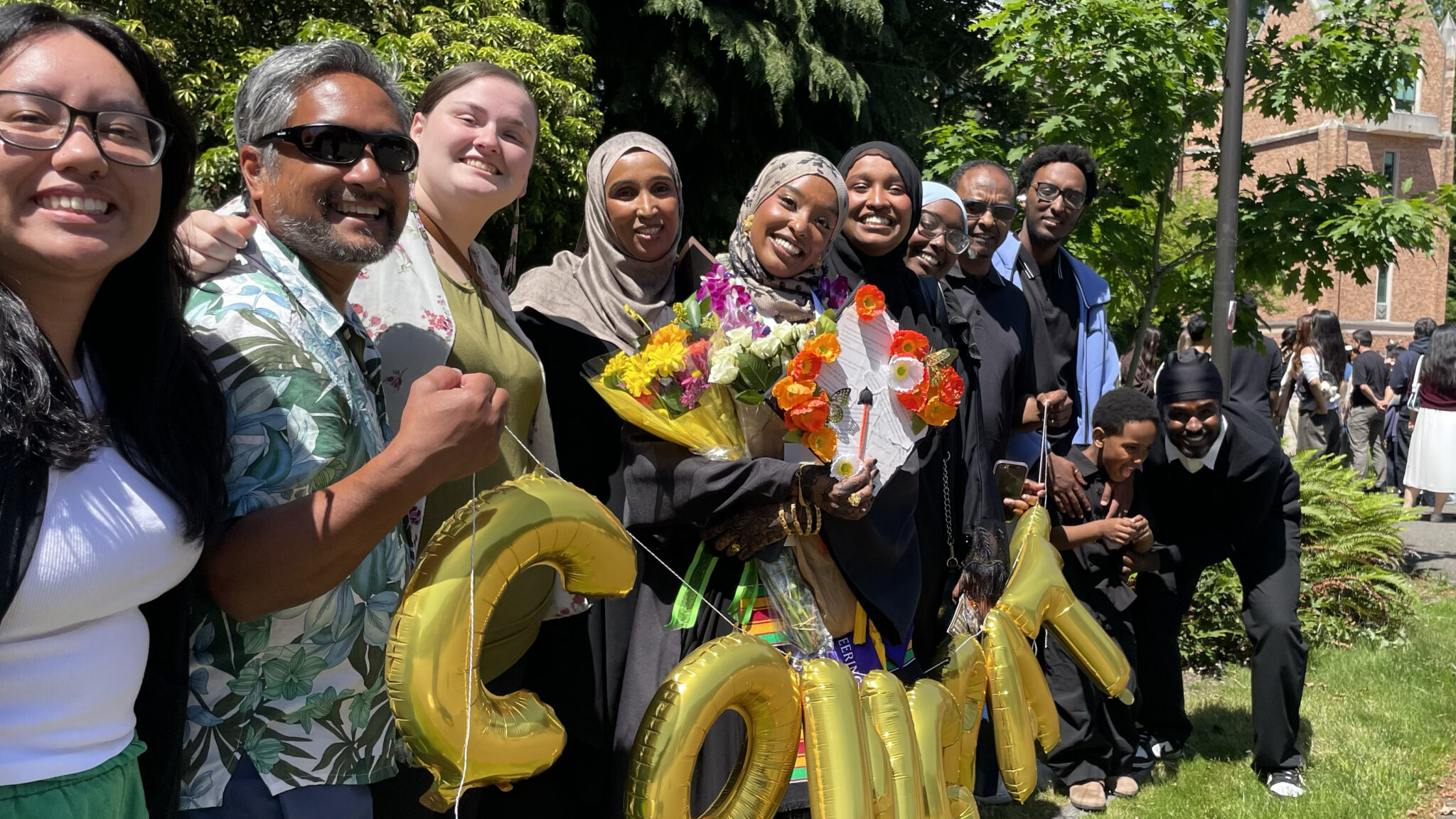 A group smiling together at a graduation ceremony. They are holding up balloons that spell "Congrats". The person in the center holds several flower bouquets.